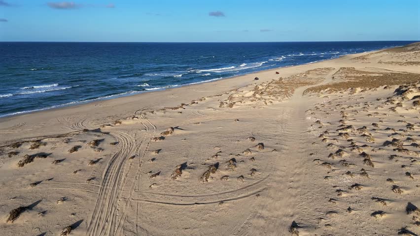 Drone descending from dunes toward the ocean in Baja California Sur, tracking two ATVs riding along the beach in warm golden hour light.