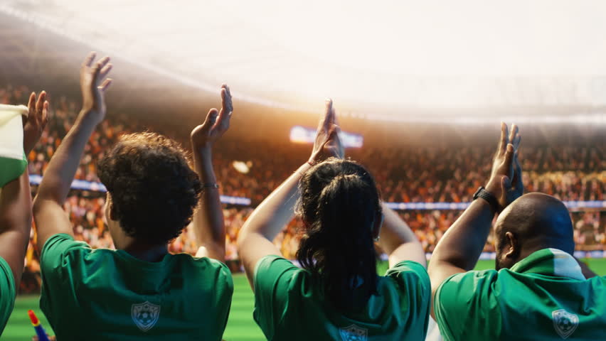 Soccer supporters cheer in packed stadium stands during a football match, creating loud crowd energy and team support, as chants and singing build match day atmosphere.