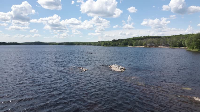 Rock formations emerging from Commanda Lake under a bright summer sky in Restoule Ontario, drone dolly over windswept water ripples