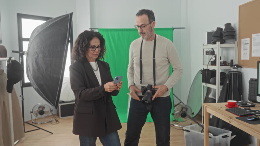 Woman and man photographers smile as woman holds smartphone and man holds camera strap while they review images in studio; creative teamwork joy.