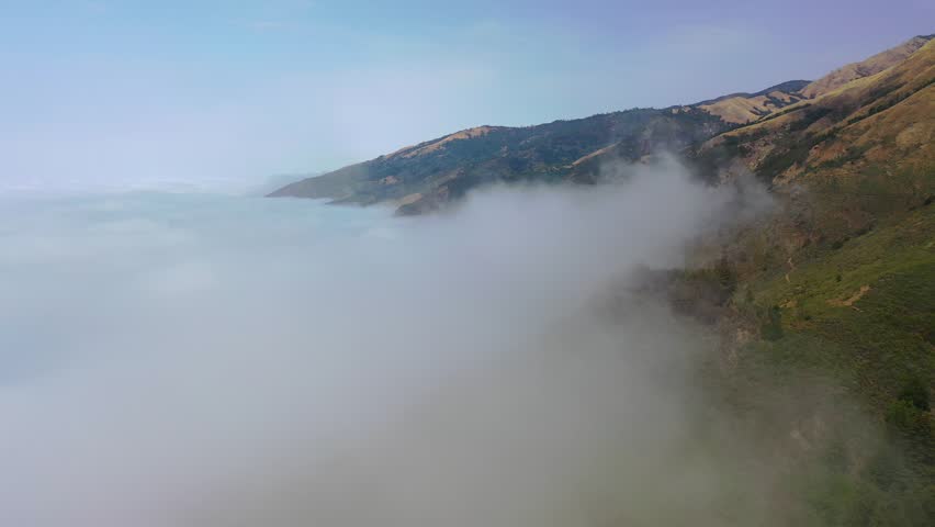Aerial drone footage flying over Big Sur mountains in the morning with a thick layer of fog and clouds over the Pacific Ocean. Blue sky overhead with sunlight on mountain ridges.