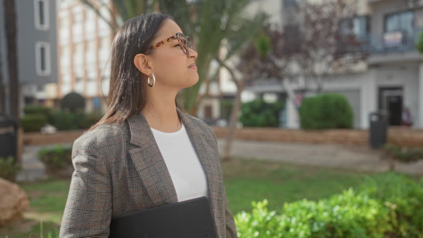 Woman in blazer holding clipboard and wearing glasses looks up smiling while standing on a street near shrubs and residential buildings; optimism.
