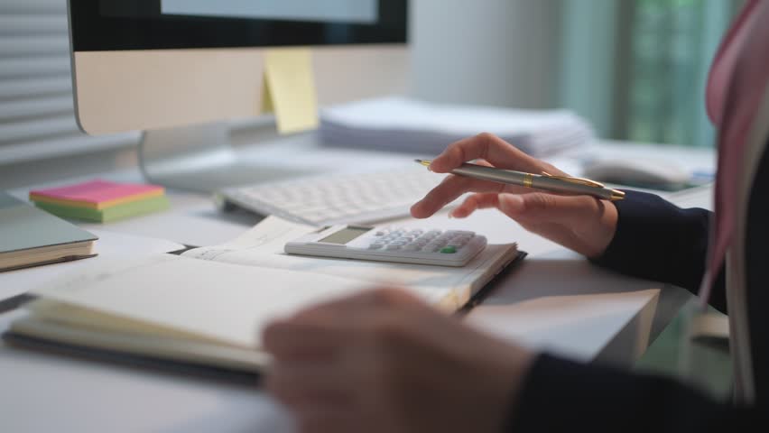Close up of a female accountant working in an office, using a calculator and writing financial data on her agenda with a pen