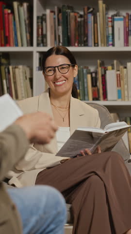 Vertical video of smiling young adult Caucasian woman with glasses holding book and listening to speaker during book presentation. Clip is perfect for promoting library events or book clubs