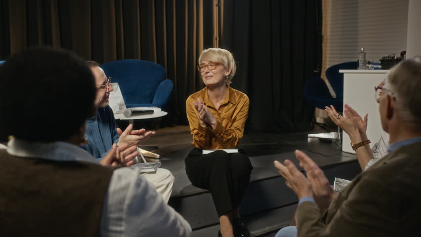 Middle-aged Caucasian woman taking off glasses and wiping tears during book presentation while audience claps. Clip is perfect for book launch promotions and author marketing campaigns, zoom in