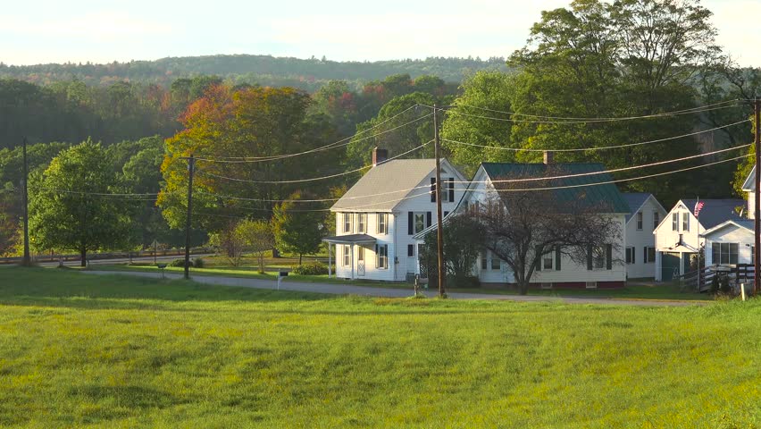 A picturesque new england autumn landscape features a classic white house, verdant fields, and a car passing on a country road with a backdrop of colorful trees and distant hills, showcasing tranquil rural life and the beauty of fall foliage in motion.