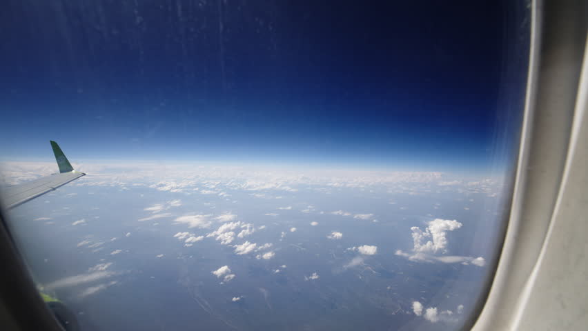 Scenic view from an airplane window showing deep blue sky, distant horizon and scattered clouds over the landscape below. Travel and aviation concept, wingtip visible at cruising altitude.