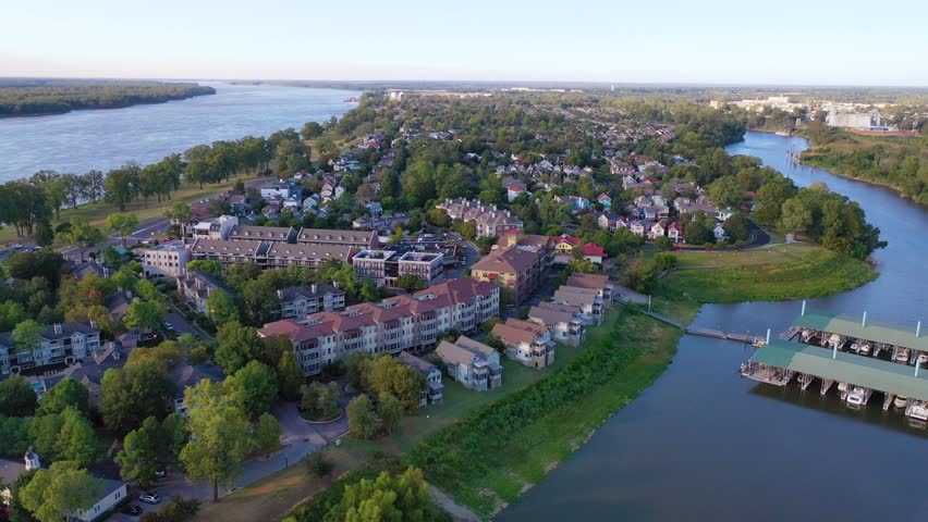Dynamic aerial footage showcases charming houses and buildings nestled along a scenic riverbank, boat docks, and lush green trees under clear blue sky, capturing serene beauty of this vibrant community.