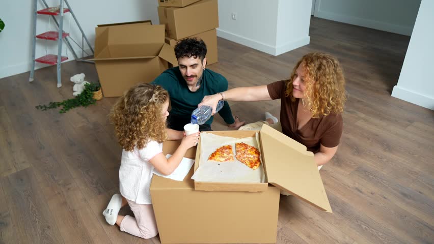 Young family taking a break from unpacking, sitting on the wooden floor amidst cardboard boxes, and joyfully sharing pizza and water to celebrate their new apartment purchase