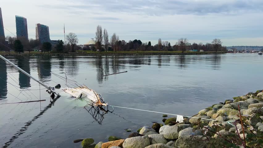 A derelict sailboat lies capsized and half-sunk beside a moss-covered breakwater at English Bay in Vancouver, its fallen mast trailing across still water. Across the calm, reflective bay the silver-domed Space Centre, leafless trees and downtown high-rise towers line the Vanier Park shore under a grey winter sky. Static footage of the wreck on the waterfront.