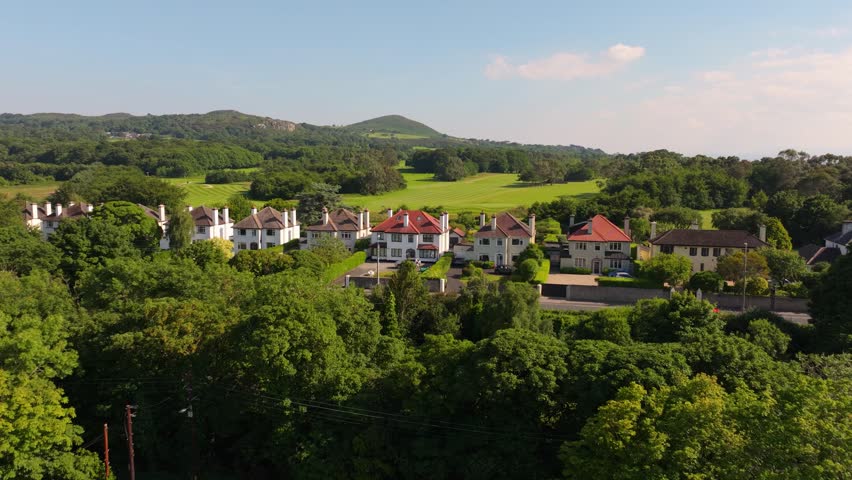 Tree-lined suburban villas front a wide green golf course and rolling hills on a clear sunny day, viewed from a drone flyover.