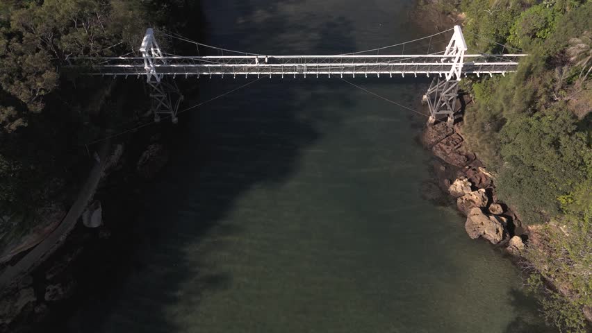 Small suspension bridge over water and bushland. Footbridge is for pedestrians crossing from one side of Parsley Bay to the other. Drone flies slowly in towards bridge as people walk across