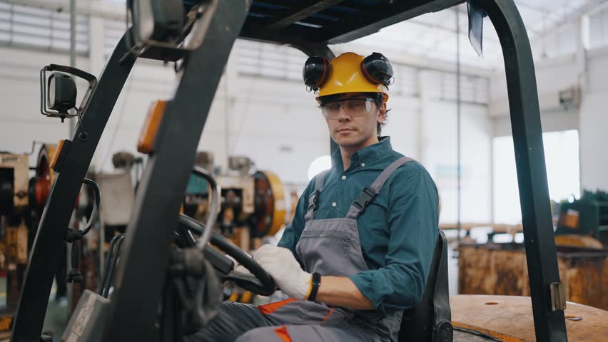 Worker stands confidently on forklift in warehouse. Wears helmet, safety glasses, and protective gloves. Background shows factory equipment and bright lighting. Forklift operator looks calm