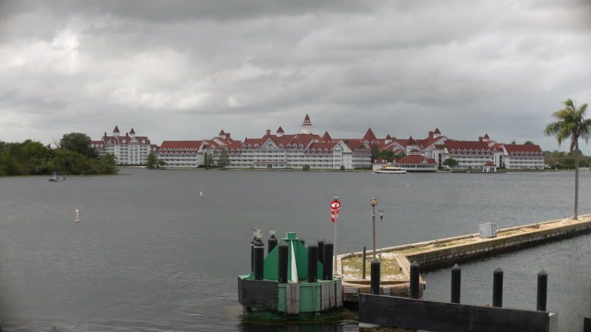 Resort hotel across a large lake is framed by docks and cloudy skies in a wide waterfront establishing shot. Grand vacation architecture, marina details, and stormy weather create a recognizable desti