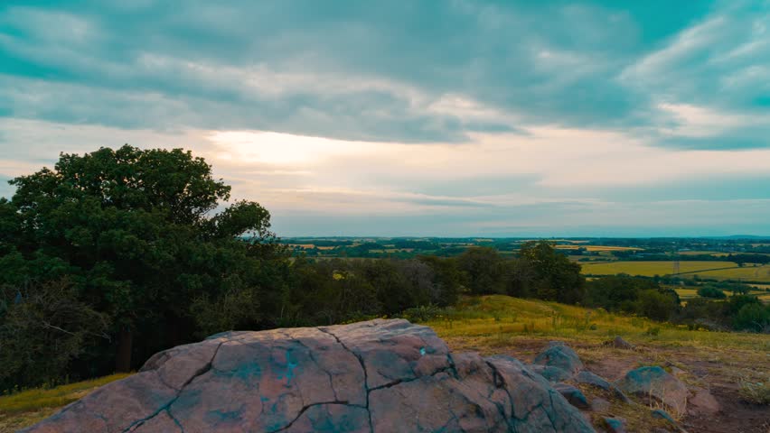 Timelapse weathered granite boulder overlooking rural countryside. Rocky hilltop viewpoint above farmland fields forest and distant agricultural horizon beneath dramatic clouds. Moving cloudscape