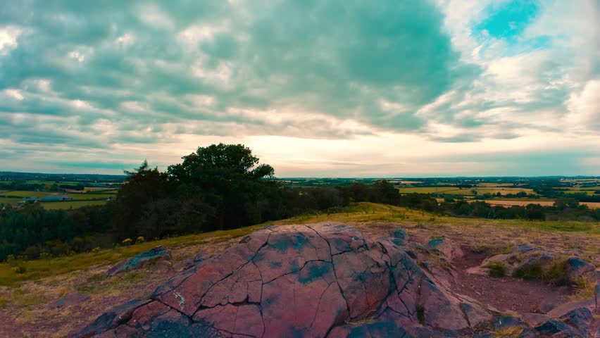 Timelapse fractured granite boulder beneath dramatic moving clouds. Hilltop hiking trail overlooking rural farmland forest and distant agricultural horizon. Panoramic countryside landscape with rocky