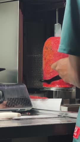 A street food vendor slicing freshly roasted kebab meat from a vertical rotisserie with a sharp knife. The skilled preparation highlights authentic Middle Eastern cuisine, showcasing juicy, flavorful meat ready to be served in wraps, sandwiches, or rice dishes