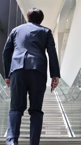 A handsome Japanese man in a suit walking up the office stairs while talking on his smartphone. Conveys the image of a mid-level business executive excelling in sales. A video showing his upper body and full body. Slow motion.