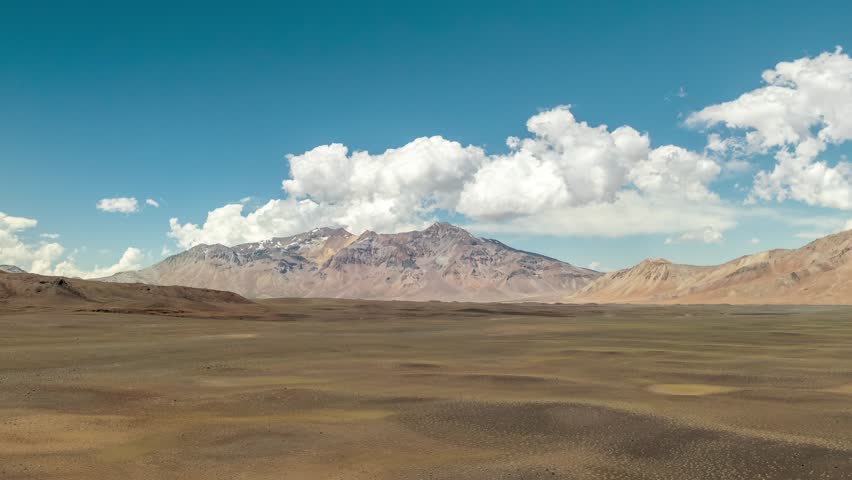A drone hyperlapse of vast desert stretches under blue sky with white clouds, framed by rugged mountains near Hito, Chile-Argentina border