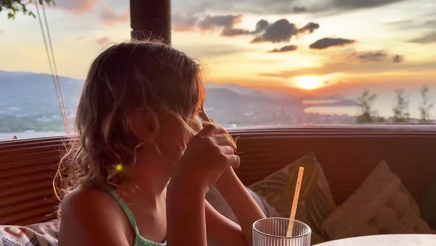 A close-up medium shot of a young girl with light brown hair, wearing a teal spaghetti-strap top, sitting on an outdoor terrace at dusk. She is looking down, drinking from a clear fluted glass with a straw, while her right hand is running through her hair. The shot is backlit by a dramatic golden sunset, casting a warm glow and significant lens flare. The background features a beautiful coastal view with mountains and patterned cushions. The image captures themes of relaxation, vacation, family moments, childhood, lifestyle, and the serene beauty of the golden hour.