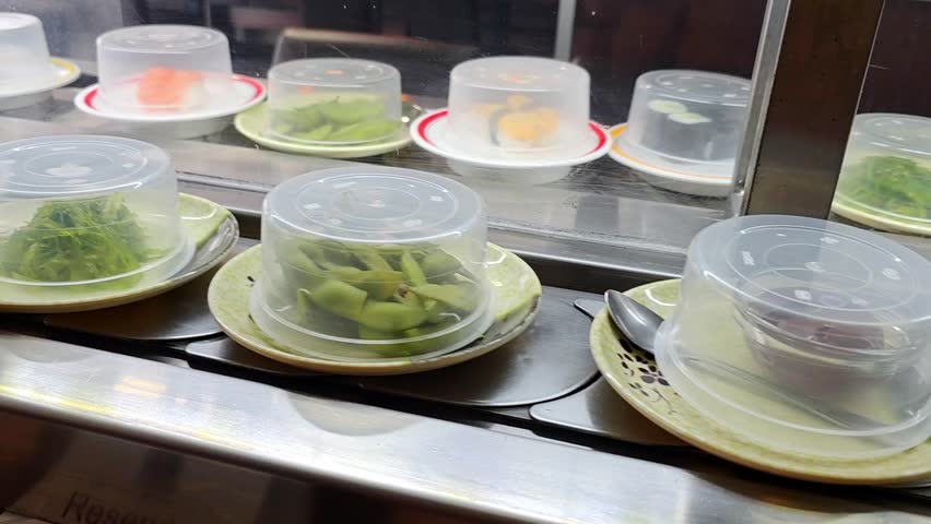 Rows of covered sushi plates move along a conveyor belt in a Japanese restaurant, varied colored dishes lined up, kaiten sushi dining, self service concept.