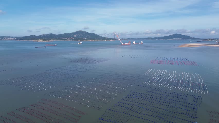 Aerial drone forward flight over vast offshore aquaculture at Pingtan Island, China. Expansive white, blue and red buoy grids stretching across calm sea. Golden tidal sandbar, mountain islands and cargo ships under bright blue sky. Epic panoramic coastal seascape with immense scale.