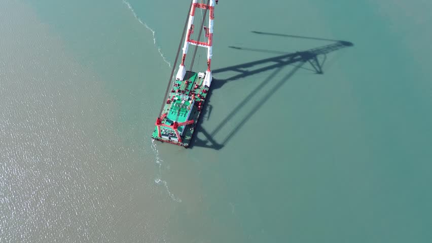 High overhead top-down aerial drone shot of 2200-ton floating crane vessel at Pingtan tidal waters, Fujian China. Vertical red-white segmented crane boom with golden truss top extends upward, multiple groups of steel cables and lifting hooks form neat vertical lines. A long clear shadow of the crane stretches diagonally on two-tone seawater, no extra vessels or mountains in frame, ultra-clean minimalist composition highlighting the precise vertical geometric structure of marine heavy-lift equipment.