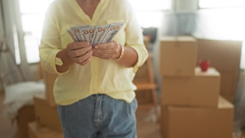 Woman counting stacks of dollars with visible hands and a smile amid moving boxes and a ladder inside a building; savings relief.