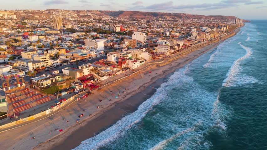 A sweeping golden hour drone shot capturing the rolling Pacific surf meeting the urban coast of Playas de Tijuana, Mexico.