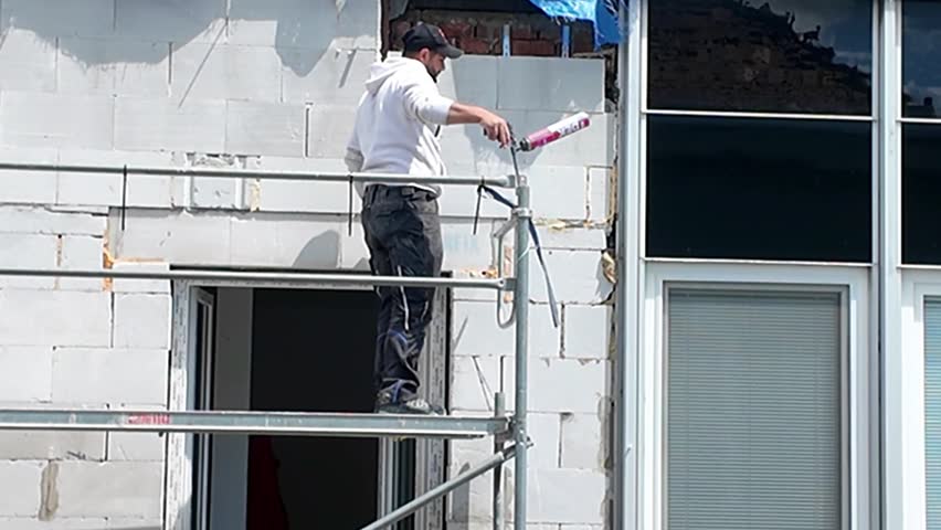 Construction worker installing windows on scaffolding at a building site. A worker in protective gear uses tools to install window frames on a building under construction, seen from scaffolding.