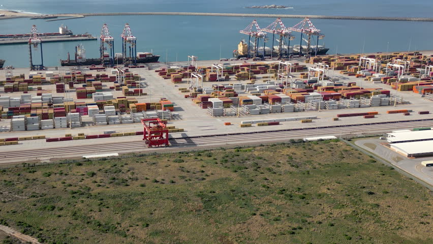 Industrial container shipping port with cranes, stacked cargo containers, rail lines and calm blue harbor viewed from above.