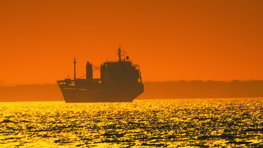 A large ship is seen sailing on the water while the sun sets in the background. The sky turns orange and the water reflects the colors of the sunset
