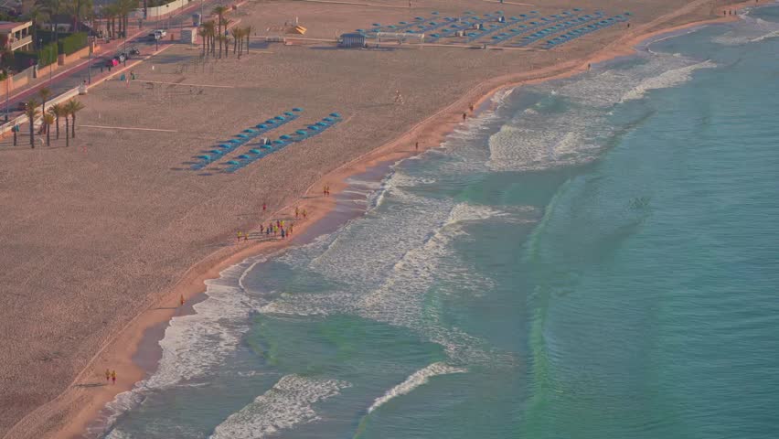 Footprints line the shore as people walk along the beach. Sun loungers are set up on the sand. Waves crash gently on the beach. The sun shines brightly on the water.