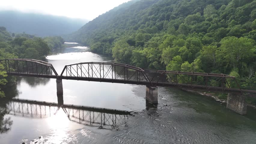 Wide drone shot of a historic metal arch bridge crossing over the wide, sunlit river winding through the forested canyon of New River Gorge National Park, West Virginia USA