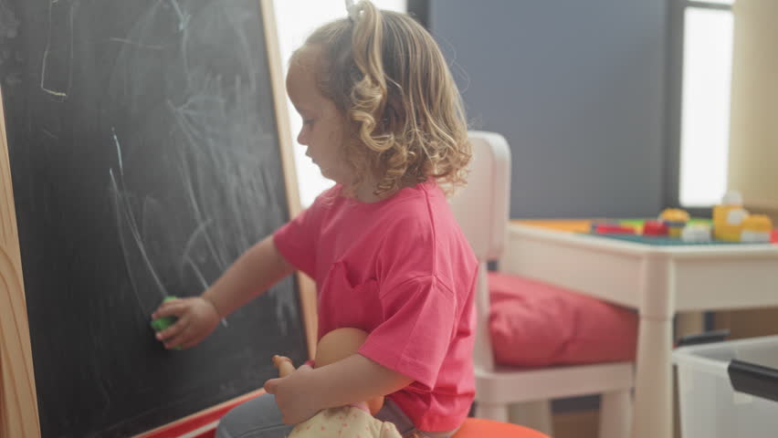 Girl erases chalkboard with green eraser while holding a doll in a playroom; curiosity play learning.
