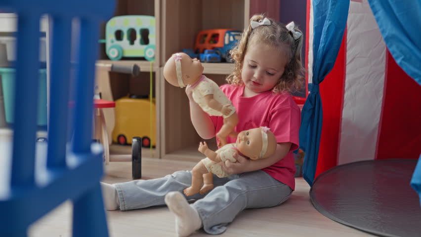 Little girl holding baby doll kneels by toy shelf in playroom building; playfulness curiosity imagination.