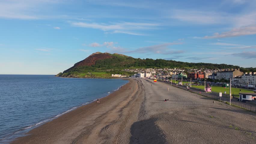 Wide coastal strand curves toward a grassy park and headland under clear summer skies with a few walkers along the shore.