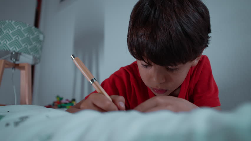 Young boy pauses during writing activity and looks away thoughtfully while lying on his bed, authentic childhood moment expressing curiosity, reflection, imagination, concentration and everyday learning at home