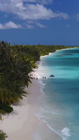 Aerial view of Dhigurah Island, Maldives, with palm lined shore, white sand, turquoise lagoon, two cell towers, gentle waves, people wading, and a jet ski at noon