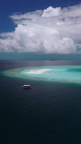 Aerial view of Dhigurah, Maldives, with a crescent sandbank, three beach tents, a drifting wooden dhoni, vivid turquoise lagoon, deep navy ocean, and cumulus clouds.
