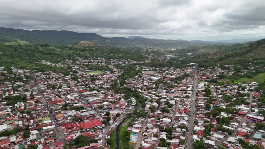 Aerial view shows houses along streets and green hills. Clouds cover the sky. The town layout is clear with roads and buildings.