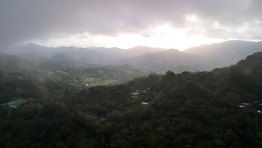 Clouds roll over hills as the sun rises. Green trees and fields stretch below. Nature surrounds the viewer in this early morning setting.