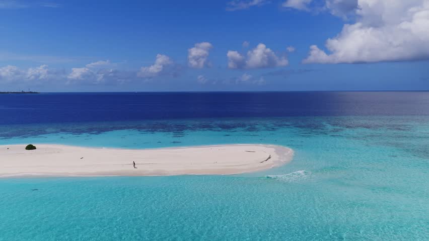 Aerial view of a crescent sandbar off Dhigurah in the Maldives, with atolls and island towers on the horizon, a person walking, a drifting boat, and gentle wave motion in midday light.