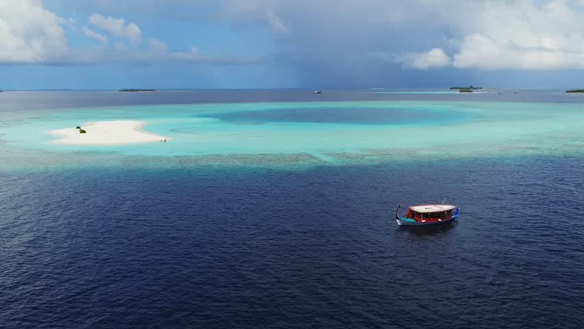 Aerial daytime view of Dhigurah island, Maldives. A dhoni drifts, visitors walk a crescent sandbank, coral reef flats encircle, a dark lagoon pool sits, low clouds line the horizon.