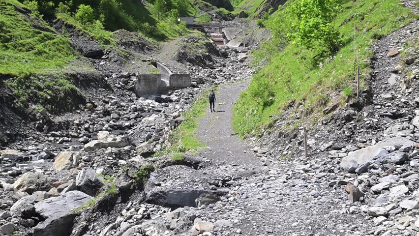 Summer mountain ravine with gravel streambed, hiking path, and green slopes beneath steep cliffs in the Swiss Alps.