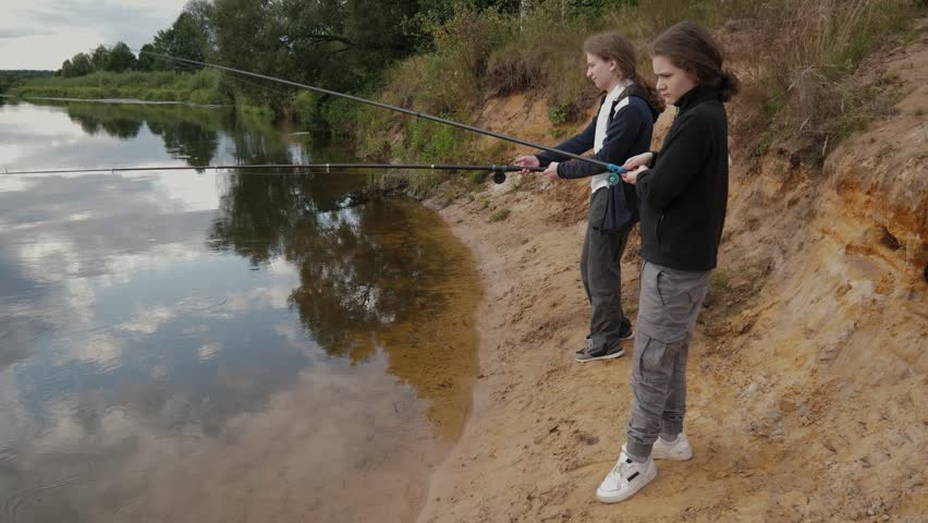 Side view of two teens standing on a sandy riverbank casting fishing lines into a calm tree-lined river under an overcast sky