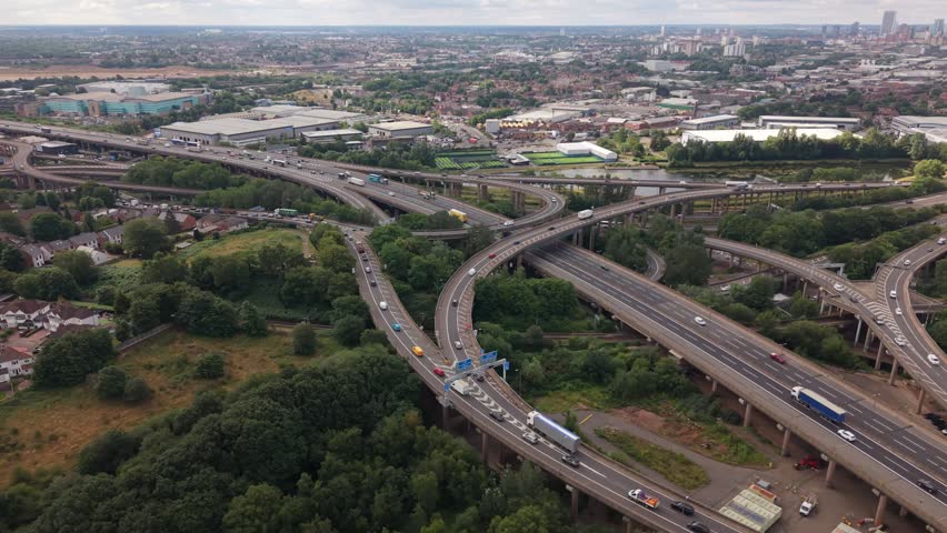 Smooth side drone flight across the iconic Spaghetti Junction motorway interchange in Birmingham, England. As the drone moves sideways, the intricate network of elevated M6 motorways and flyovers gives way to a panoramic view of Birmingham city skyline in the distance. Cinematic aerial footage featuring urban infrastructure, heavy traffic and the West Midlands landscape.