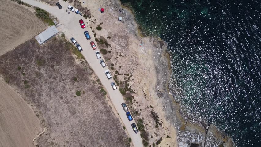 Gozo, Malta, L-Ahmar salt pans. stationary vehicles along the coastal road with aerial view of sandy terrain and water body. neutral, observational perspective on real estate, transportation. High