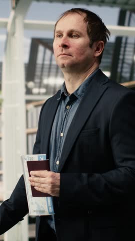 Portrait format video: Man holding passport and trolley handle in airport terminal, catching flight, tourist carrying travel documents and luggage while going to airplane departing gate.