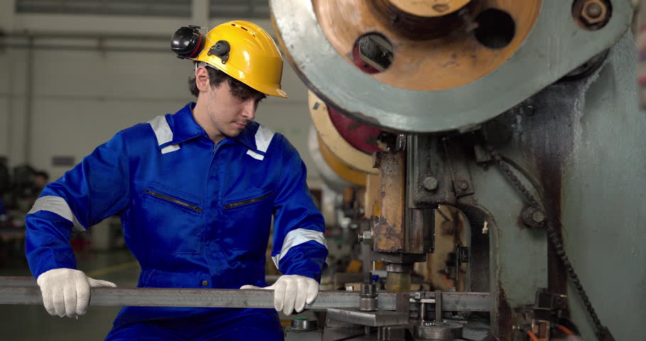 A factory worker in a blue coverall, hard hat and gloves operates a mechanical power press machine, feeding a metal bar to stamp parts on a production line in an industrial plant.